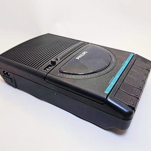 Op de afbeelding: Zwarte Philips cassettespeler met een geribbelde bovenkant en een rond bedieningspaneel. Het apparaat heeft een reeks rechthoekige knoppen aan de rechterkant en een turquoise accentstrip. Het Philips-logo is zichtbaar.