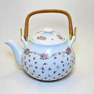 Peut inclure: Théière en céramique blanche ornée de motifs floraux roses et de feuilles vertes. La théière a un bec verseur incurvé, un couvercle avec un bouton et une anse en rotin tressé. Article de cuisine décoratif.