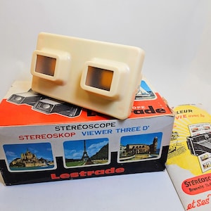 Puede incluir: Un visor de estereoscopio vintage de color crema con dos lentes, apoyado en una caja con el texto "STEREOSCOPE VIEWER THREE D". La caja presenta imágenes de monumentos y la marca "Lestrade". También hay un folleto.