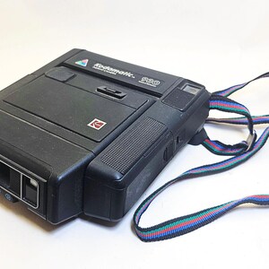 Op de afbeelding: Een zwarte Kodak Instamatic 360 camera met een veelkleurige gestreepte riem. De camera heeft een rechthoekige vorm met een lens aan de voorkant en het Kodak-logo. De camera staat op een witte ondergrond.