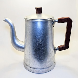 Peut inclure: Une cafetière vintage de couleur argentée avec une poignée et un bouton de couvercle en bois brun foncé. La cafetière a un bec verseur incurvé et un corps cylindrique. La poignée est rectangulaire et fixée sur le côté.