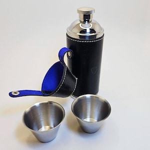 May include: A black leather flask with a silver top and two stainless steel shot glasses. The flask has a blue leather carrying case with a handle. The flask is approximately 20 cm tall.