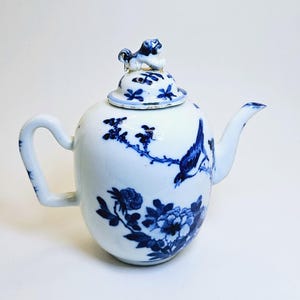 Op de afbeelding: Witte keramische theepot met blauwe bloemen- en vogelontwerpen. De theepot heeft een gebogen handvat, een tuit en een deksel met een decoratieve knop. De blauwe ontwerpen omvatten bloemen, takken en een vogel.
