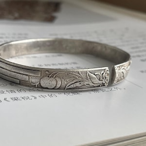 May include: A silver-toned open cuff bracelet featuring a textured surface with embossed botanical motifs, including rounded fruit-like shapes and leaf patterns, alongside a series of parallel horizontal lines on the outer band.