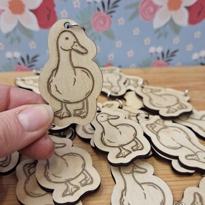 May include: A collection of wooden duck keyrings, each featuring a detailed outline of a duck. The keyrings are made from light-coloured wood and have a small metal ring. One keyring is held by a hand, with others scattered around.