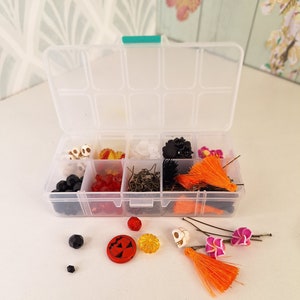 Puede incluir: Una caja de almacenamiento de plástico transparente llena de diversos materiales de artesanía con temática de Halloween, que incluyen cuentas negras, cuentas rojas y naranjas, dijes de calavera naranja y blanca, dijes de flores naranja y blanca, borlas naranjas y hallazgos metálicos dorados. La caja está abierta y algunos de los materiales están esparcidos sobre una superficie blanca.