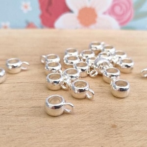 May include: A group of silver-toned metal bead caps with a small loop on one side. The caps are scattered on a light brown wooden surface.
