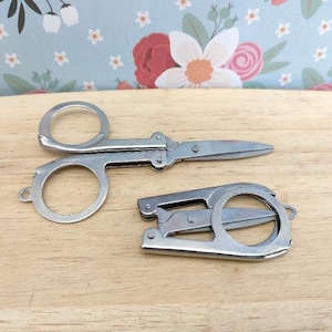 May include: Two silver folding scissors with a ring handle. The scissors are on a light brown wooden surface.