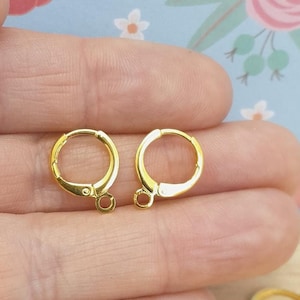 May include: Two gold-toned earring hoops with a hinged closure. The hoops are shown on a hand.