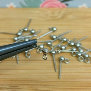 May include: A close-up of silver earring findings with a loop on the end. The findings are scattered on a wooden surface. A pair of pliers is holding one of the findings.
