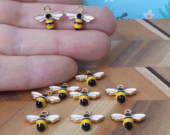 May include: Ten gold bee charms with black and yellow stripes and white wings. The charms are on a wooden surface.