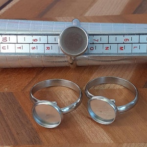 Może przedstawiać: Dwa srebrne blankiety pierścionków z okrągłymi obramowaniami. Obrąbki mają średnicę około 2,5 cm. Pierścienie znajdują się na metalowym mierniku rozmiaru pierścionków.
