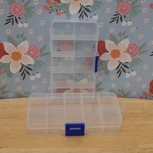 May include: A clear plastic storage box with multiple compartments. The box has a blue latch on the front and is open to reveal the compartments inside.