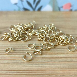 May include: A scattering of small, gold-coloured open jump rings on a light brown wooden surface. The rings are in various orientations, some clustered together and others isolated. The lighting highlights the metallic sheen of the jewellery findings.