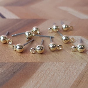 May include: Eight gold-toned earring findings with a small loop on one end and a ball on the other end. The findings are arranged on a wooden surface.