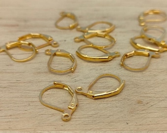 May include: A group of 10 gold-toned earring hooks, also known as earring findings, are scattered on a light wood surface. The hooks are made of metal and have a spring-loaded clasp.