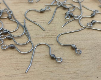 May include: A collection of silver-toned metal earring hooks, each featuring a curved wire ear wire, a small coiled spring detail, and a circular loop at the bottom for attaching jewelry components, scattered across a light-colored wooden surface.