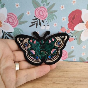 May include: An embroidered butterfly patch in green and black with pink and blue accents. The patch is on a wooden surface.