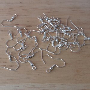 Puede incluir: Un mont&oacute;n de ganchos para pendientes de plata con peque&ntilde;as bolas redondas en la parte superior. Los ganchos est&aacute;n esparcidos sobre una superficie de madera.