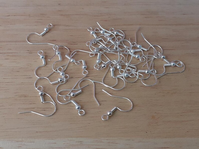 Puede incluir: Un mont&oacute;n de ganchos para pendientes de plata con peque&ntilde;as bolas redondas en la parte superior. Los ganchos est&aacute;n esparcidos sobre una superficie de madera.