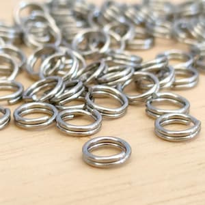 May include: A close-up of numerous small, silver-coloured double split rings. The metal rings are scattered on a light brown surface. The rings are made of a shiny, metallic material and are designed to be durable.