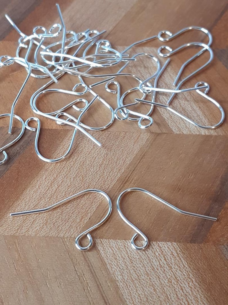 May include: A pile of silver earring hooks, a type of jewelry finding, on a wooden surface. The hooks are made of metal and have a simple design.