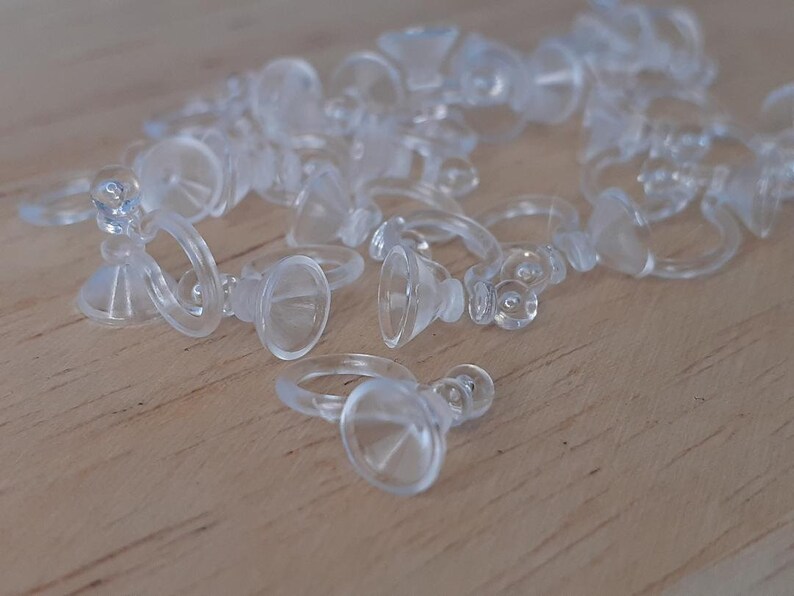 May include: A group of clear plastic suction cup hooks with a ring on the top. The hooks are scattered on a light wood surface.