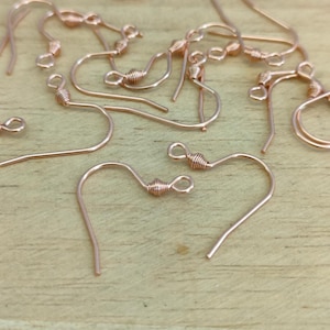 May include: A close-up of a group of rose gold earring hooks. The hooks are made of metal and have a small loop at the top. They are scattered on a wooden surface.