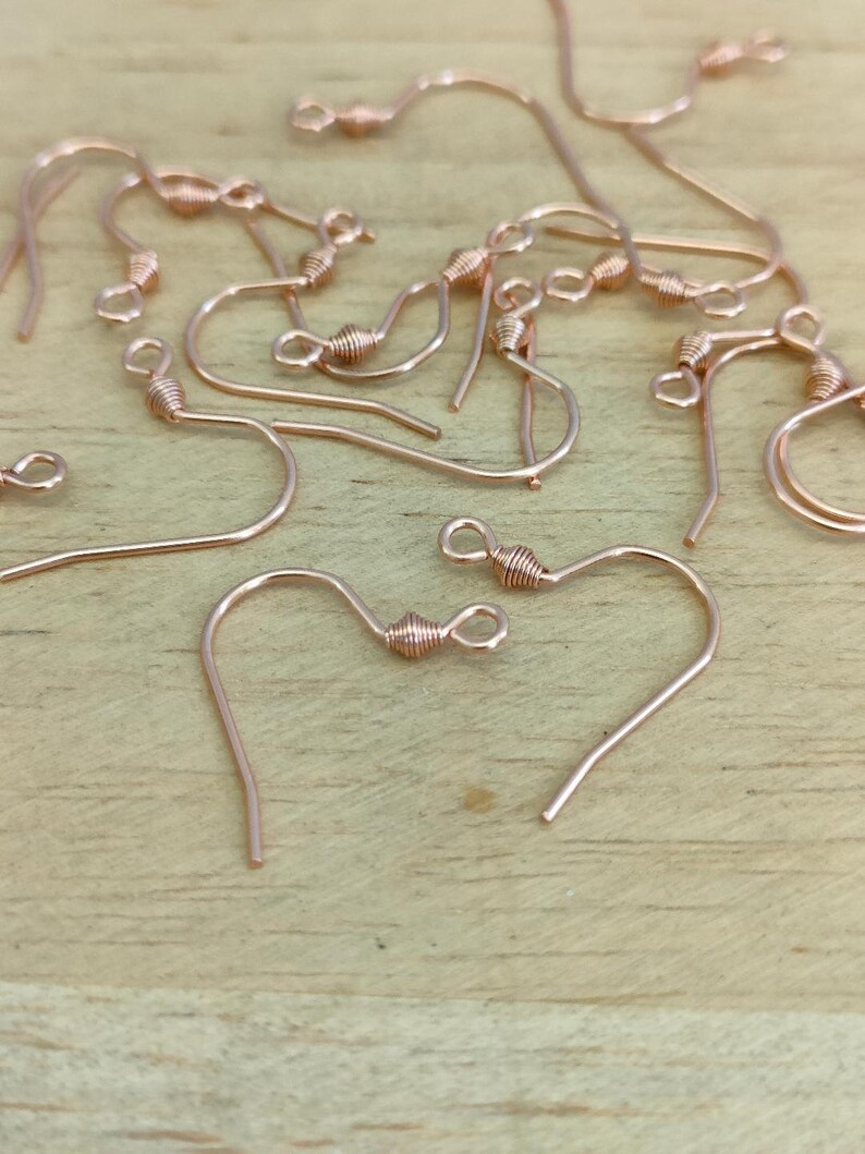 May include: A close-up of a group of rose gold earring hooks. The hooks are made of metal and have a small loop at the top. They are scattered on a wooden surface.