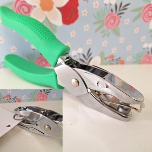 May include: A green handled heart shaped paper punch with a silver metal punch head. The punch is on a white surface with a floral pattern in the background.