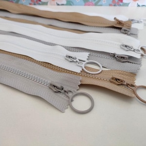 May include: A close-up of a set of five zipper pulls in various shades of beige and grey. The zipper pulls are arranged in a stack, with the top pull being the lightest colour and the bottom pull being the darkest colour. Each pull has a small metal ring attached to it.