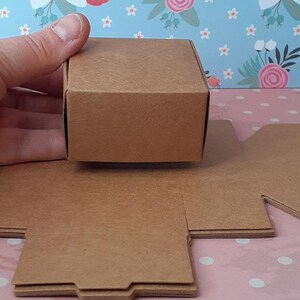 K&ouml;nnte beinhalten: Ein Satz brauner Pappkartons, flach gefaltet, mit einem zusammengebauten Karton, der in einer Hand gehalten wird. Die Kartons sind zum Verpacken von kleinen Geschenken oder Gef&auml;lligkeiten gedacht.