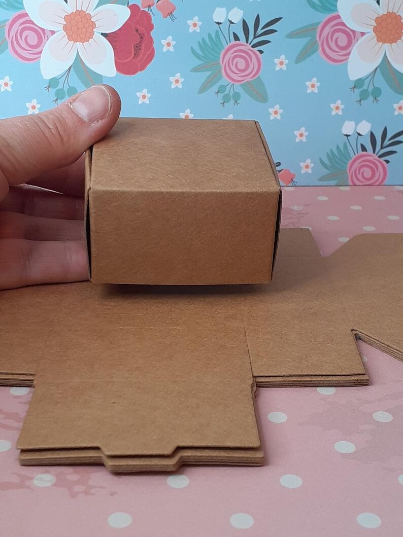K&ouml;nnte beinhalten: Ein Satz brauner Pappkartons, flach gefaltet, mit einem zusammengebauten Karton, der in einer Hand gehalten wird. Die Kartons sind zum Verpacken von kleinen Geschenken oder Gef&auml;lligkeiten gedacht.