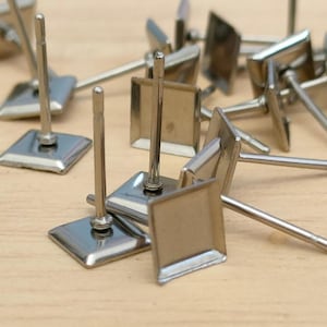 May include: A close-up of a pile of silver earring posts with square bases. The posts are made of metal and have a shiny finish.