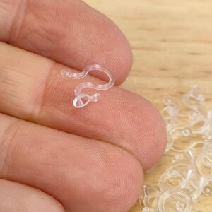 May include: A close-up of a clear plastic hook, with a pile of similar hooks in the background. The hook is designed to hold items together.