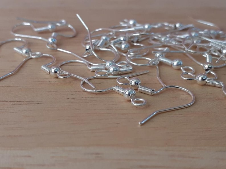 Puede incluir: Primer plano de un mont&oacute;n de ganchos para pendientes de plata. Los ganchos son de metal y tienen una peque&ntilde;a bola en la parte superior. Se utilizan com&uacute;nmente para hacer pendientes.