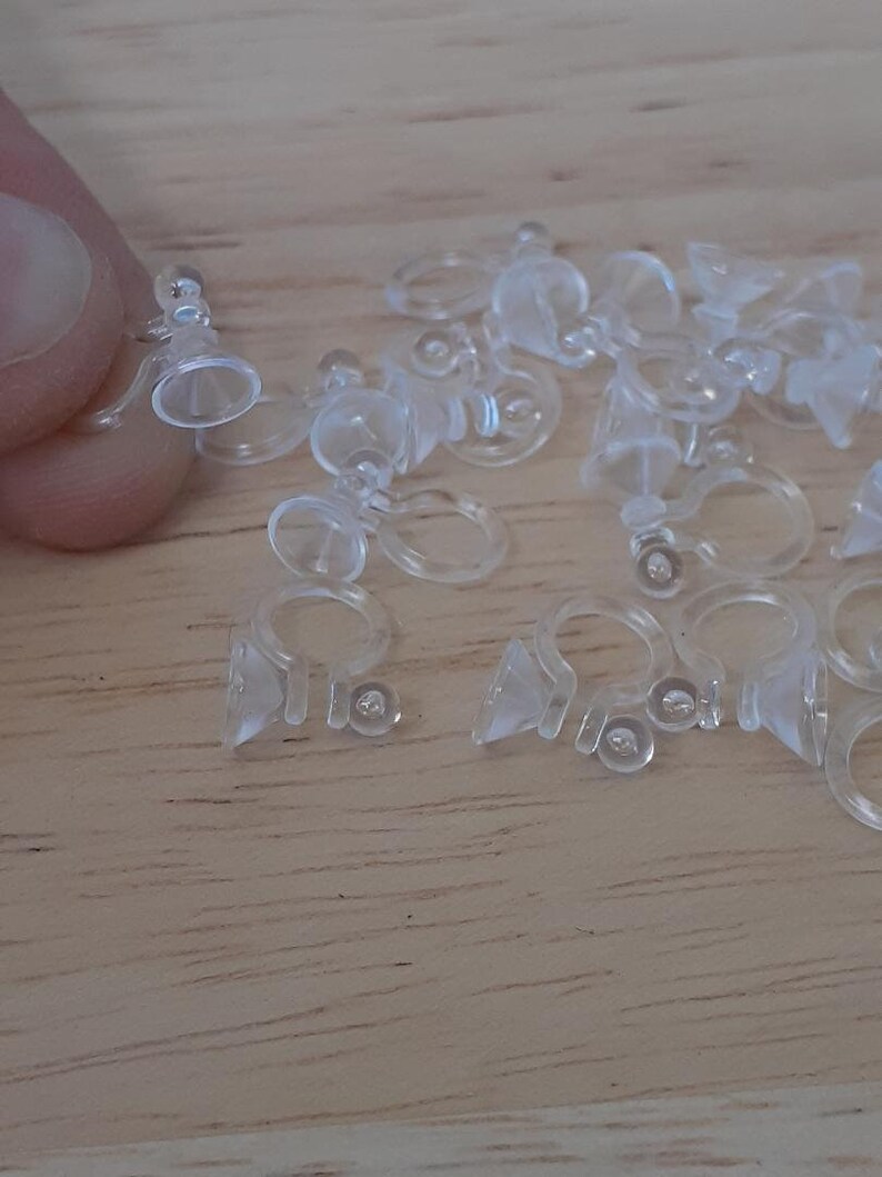 May include: A close-up of a pile of clear plastic earring backs. The backs are designed to be used with post earrings and have a small suction cup on the bottom to help keep them in place.