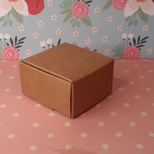 K&ouml;nnte beinhalten: Eine kleine, braune Pappbox mit quadratischer Form. Die Box steht auf einer rosa Oberfl&auml;che mit wei&szlig;en Punkten.