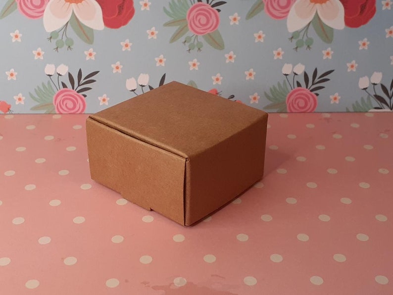 K&ouml;nnte beinhalten: Eine kleine, braune Pappbox mit quadratischer Form. Die Box steht auf einer rosa Oberfl&auml;che mit wei&szlig;en Punkten.
