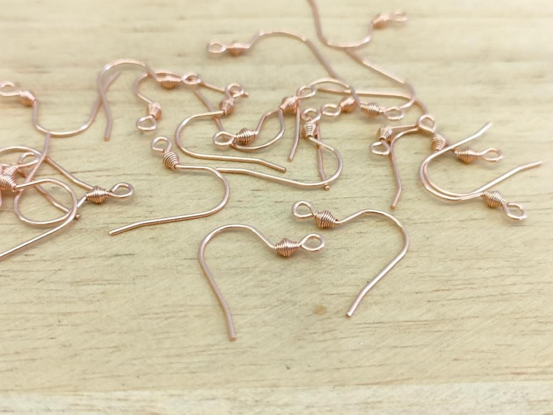 May include: A close-up of a pile of rose gold earring hooks. The hooks are made of wire and have a small loop at the top. The hooks are all twisted at the top to create a decorative effect.