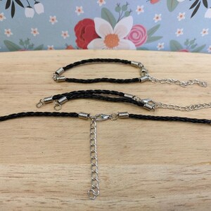 May include: Three black braided cords with silver metal clasps. The cords are arranged in a line with the top cord being the shortest and the bottom cord being the longest. The cords are on a wooden surface.