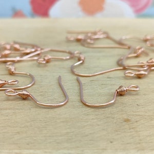 May include: A close-up of a group of rose gold earring hooks. The hooks are made of wire and have a small loop at the top. The hooks are arranged in a random pattern on a wooden surface.