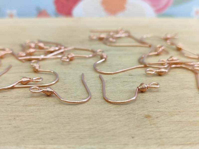 May include: A close-up of a group of rose gold earring hooks. The hooks are made of wire and have a small loop at the top. The hooks are arranged in a random pattern on a wooden surface.