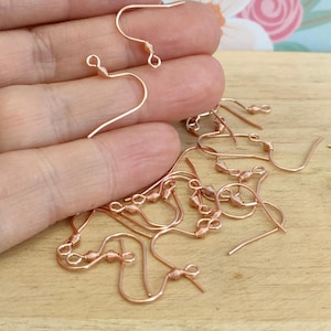 May include: A close-up of a pile of rose gold earring hooks. The hooks are made of wire and have a small loop at the top. The hooks are designed for making earrings.