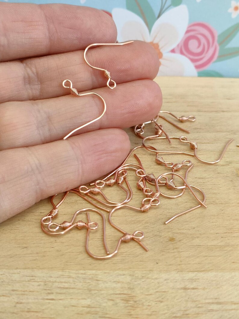 May include: A close-up of a pile of rose gold earring hooks. The hooks are made of wire and have a small loop at the top. The hooks are designed for making earrings.