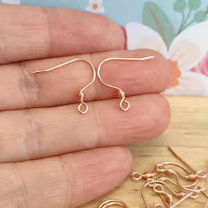 May include: Rose gold earring hooks, a close-up of two hooks held in a hand, and a pile of additional hooks on a wooden surface.