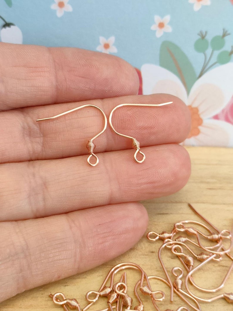 May include: Rose gold earring hooks, a close-up of two hooks held in a hand, and a pile of additional hooks on a wooden surface.