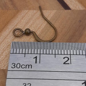 Może przedstawiać: Zbliżenie na złoty haczyk do kolczyk&oacute;w z małą kulką na g&oacute;rze. Haczyka zmierzono linijką, co pokazuje, że ma około 1 cm długości.
