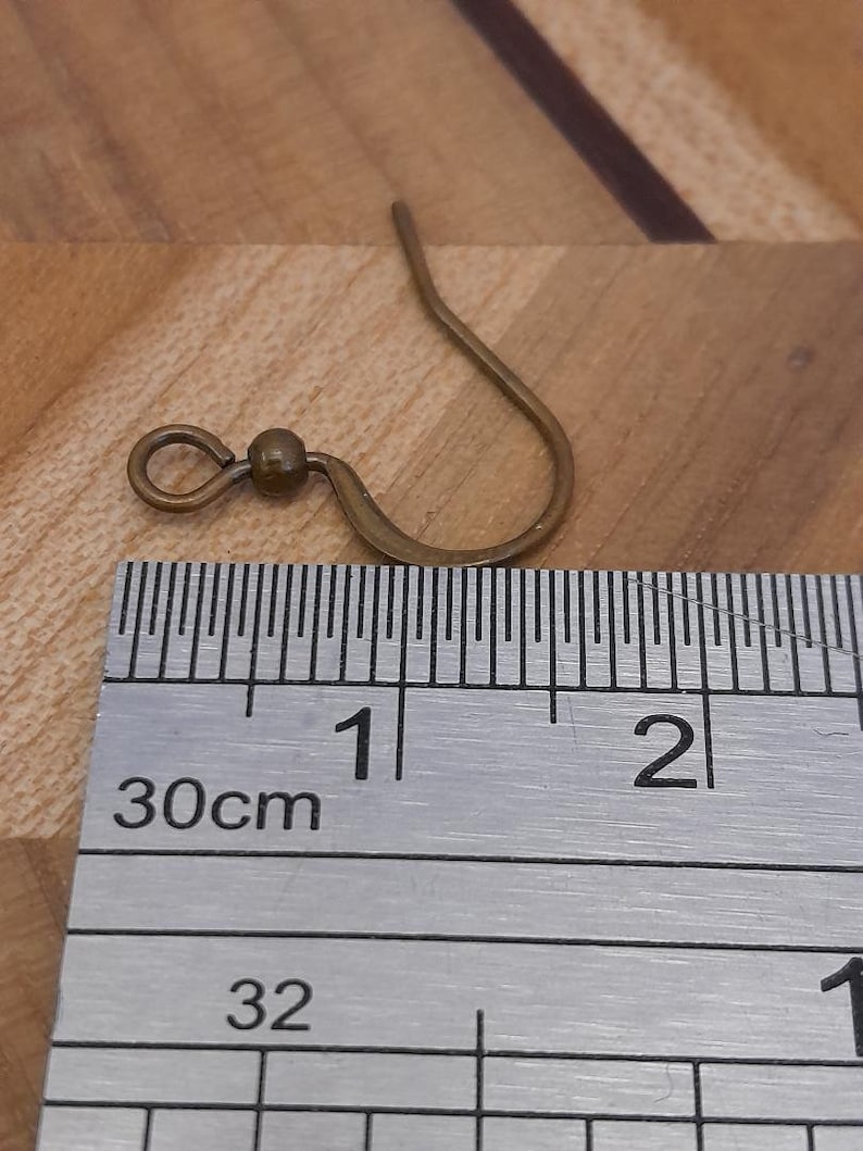 Może przedstawiać: Zbliżenie na złoty haczyk do kolczyk&oacute;w z małą kulką na g&oacute;rze. Haczyka zmierzono linijką, co pokazuje, że ma około 1 cm długości.