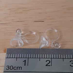 May include: Two clear plastic earring backs with a suction cup on the bottom. The earring backs are measured against a ruler, showing they are about 1 cm in length.